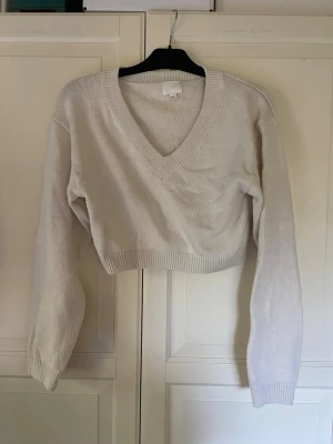 Krämvit croppad stickad tröja - Snygg croppad stickad tröja i krämvit med djup v-ringning och långa ärmar. Tröjan har ribbade muddar vid ärmslut och nederkant, vilket ger en stilren och modern look. Perfekt att matcha med höga jeans eller kjol.