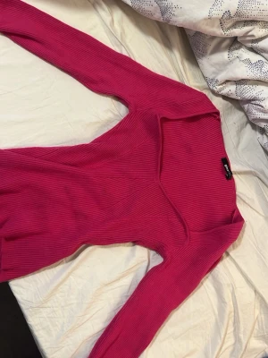 Ribbad rosa topp med v-ringning - Trendig ribbad topp i starkt rosa färg med lång ärm och djup v-ringning. Figurnära passform och stretchigt material som ger en snygg siluett. Perfekt för att lyfta vilken outfit som helst.