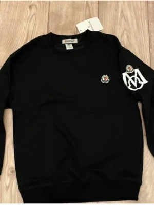 Svart sweatshirt med logga - Svart sweatshirt med rund hals och långa ärmar. Tröjan har en liten broderad logga på bröstet och en större vit tryckt detalj på ena ärmen. Perfekt för en avslappnad och trendig stil.