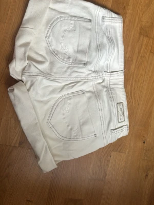 Vita denimshorts med dubbla knappar - Säljer ett par vita denimshorts med dubbla knappar framtill och klassiska fem fickor. Shortsen har upprullade benslut och kontrastsömmar. Perfekta för varma dagar och ger en fräsch look till din outfit.