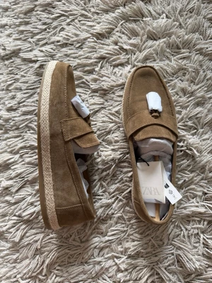 Beige mockaloafers med flätad sula - Snygga loafers i beige mocka med klassisk penny strap och en flätad, espadrille-inspirerad sula. Skorna har en stilren och avslappnad look som passar perfekt till jeans eller chinos. Märket är Zara och de har en låg profil och rundad tå.