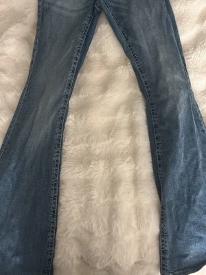 Blå bootcut med broderade fickor - Snygga blå bootcut jeans med slitningar och broderade detaljer på bakfickorna. Jeansen har en klassisk femficksmodell och är tillverkade i denim med lätt tvättad look. Perfekta för dig som gillar en retro och avslappnad stil.