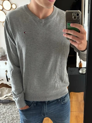 Grå V-ringad tröja Tommy Hilfiger - Snygg grå V-ringad tröja från Tommy Hilfiger med liten broderad logga på bröstet. Tröjan har långa ärmar, ribbade muddar och är tillverkad i mjukt material som passar perfekt till jeans eller chinos.