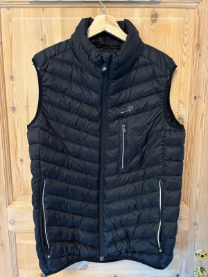 Svart dunväst från Pelle P - Stilren svart dunväst från Pelle P med hög krage och tre dragkedjeförsedda fickor. Västen har quiltad design och diskret logga på bröstet. Perfekt för lager-på-lager och håller dig varm under kyliga dagar.