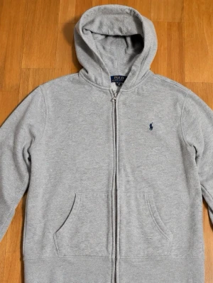 Grå Ralph Lauren hoodie - Klassisk grå hoodie med dragkedja från Polo Ralph Lauren. Tröjan har huva, två fickor framtill och den ikoniska mörkblå loggan broderad på bröstet. Tillverkad i mjukt sweatshirtmaterial som är perfekt för chill dagar. Den är STRL XS om du undrade