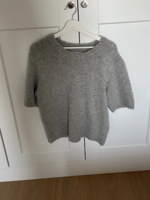 Grå stickad tröja med kort ärm - Mysig grå stickad tröja med korta ärmar och rund hals. Tröjan har en fluffig och mjuk struktur, perfekt för lager på lager. Passar till både jeans och kjol för en avslappnad look. Lite oversized men storlek S. Från samsoe samsoe nypris:700kr