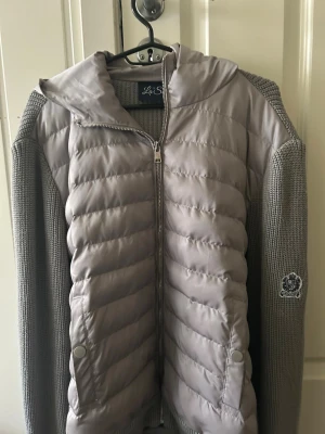 Grå cardigan jacka från La Santino - Snygg grå cardigan jacka med stickade ärmar och rygg samt quiltad framsida och huva. Jackan har dragkedja framtill, två fickor med tryckknappar och en patch på ena ärmen. Perfekt för dig som vill ha en  varm vinter