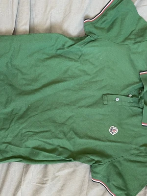 Grön pikétröja från Moncler - Snygg grön pikétröja från Moncler med klassisk krage och knappar framtill. Tröjan har Moncler-logga broderad på bröstet och vita samt röda detaljer på krage och ärmslut. Tillverkad i pikétyg som känns både sportigt och stilrent.