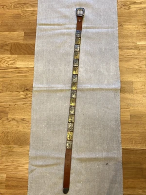 Brunt bälte med metallplattor 97cm - Unikt brunt bälte i läder med dekorativa fyrkantiga metallplattor i silver och guldtoner längs hela remmen. Spännet är i metall och har en klassisk form. Perfekt accessoar för att lyfta din outfit med en cool vintagekänsla.