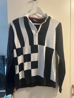 Svartvit långärmad polotopp med mönster - Snygg långärmad polotopp i svart och vitt med grafiskt rutigt och randigt mönster framtill. Krage i vitt och djup v-ringning. Perfekt för dig som vill sticka ut med en unik stil. Materialet känns mjukt och bekvämt.