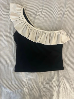 Svart topp med volang från H&M - Snygg svart topp från H&M med en axel och stor vit volang längs halsringningen. Toppen är figurnära och har en trendig asymmetrisk design som sticker ut. Perfekt för dig som gillar unika detaljer och stilren look.