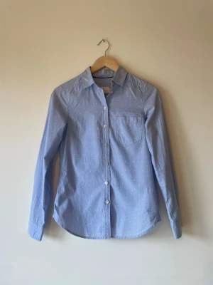 Ljusblå skjorta med prickigt mönster - Snygg ljusblå skjorta med vita prickar och klassisk krage. Skjortan har långa ärmar, bröstficka och knäppning framtill. Perfekt till jeans eller kjol för en clean och fräsch look.