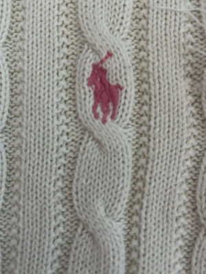 Vit kabelstickad tröja Polo Ralph Lauren - Kabelstickad vit tröja från Polo Ralph Lauren med rosa broderad logga på bröstet. Tröjan har rund halsringning och en snygg blommig detalj på insidan av kragen. Tillverkad i bomull för en mjuk och skön känsla. Någon tråd har råkat åka ut men det går att laga förmodar jag🥰 (se bild 4). Knappt använd! Vid intresse: kontakta eller klicka på köp direkt💞!