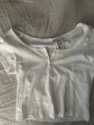 Vit croppad topp med knappar - Säljer en vit croppad topp med korta ärmar och knappdetaljer framtill. Toppen har en rund halsringning och är tillverkad i mjuk bomull, perfekt för en enkel och clean look.