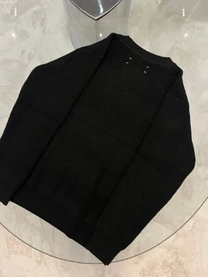 Masion margiela Paris tröja  - Snygg svart stickad tröja med rund halsringning och ribbade muddar. Tröjan har en minimalistisk design och är tillverkad i ett tjockare, mjukt material som passar perfekt till kyligare dagar. Klassisk passform och diskreta detaljer vid nacken.