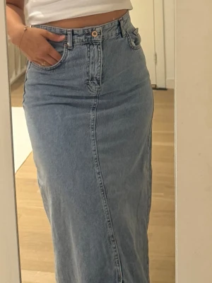 Lång jeanskjol i ljusblå denim - Säljer en lång jeanskjol i ljusblå denim med klassisk femficksdesign och hög midja. Kjolen har en rak passform och stängs med knapp och dragkedja framtill. Perfekt att styla med en crop top eller hoodie.