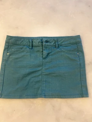 Blå jeanskjol med fickor - Snygg blå jeanskjol i kort modell med klassiska fickor fram och bak samt hällor för bälte. Kjolen har en enkel design och stängs med knapp och dragkedja framtill. Perfekt att matcha med en t-shirt eller hoodie för en chill look.