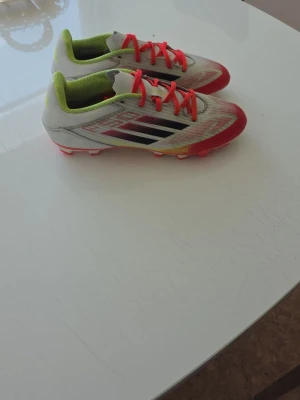Vita och röda fotbollsskor med neon - Säljer ett par snygga fotbollsskor i vitt med röda detaljer och neonfärgade insidor. Skorna har svarta ränder på sidan och röda snören. Perfekta för dig som vill sticka ut på planen. Yttermaterialet är syntet och sulan är anpassad för spel på gräs.