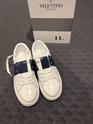 Vit och marinblå sneaker Valentino - Snygga vita sneakers från Valentino med bred marinblå rem över mitten och vita snören. Skorna har en klassisk låg siluett och är tillverkade i läder med gummisula. Perfekta för dig som gillar stilrena och exklusiva sneakers.