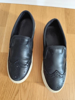 Svarta loafers i läder - Säljer ett par svarta loafers i läder med vit sula. Skorna har dekorativa perforeringar framtill och en klassisk slip-on design. Perfekta för dig som vill ha en stilren och bekväm sko till vardags. Passar till många olika outfits.
