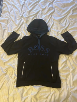 Svart hoodie från Hugo Boss - Snygg svart hoodie från Hugo Boss med stor logga på bröstet och vita detaljer vid fickorna. Hoodien har huva med dragsko och långa ärmar. Perfekt för en avslappnad och trendig stil. Använd några gånger men inga synliga fel förutom det som syns på de 2 sista bilderna som visar att det är små stygn som sticker ut men ingenting som förstör tröjans utseende 