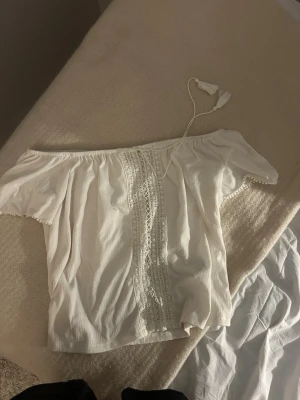 Vit off shoulder topp med spetsdetaljer - Söt vit off shoulder topp med korta ärmar och dekorativa spetsdetaljer framtill. Toppen har resår vid axlarna och en knytning upptill. Perfekt för en somrig och avslappnad stil. Lappen är bortklippt men jag vet märke och storlek💓