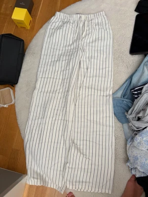 Randiga linnebyxor från H&M - Säljer ett par vita linnebyxor från H&M med tunna svarta ränder. Byxorna har rak passform, resår i midjan och är tillverkade i ett luftigt linnematerial. Perfekta för en avslappnad och stilren look.