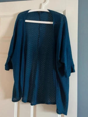 Blå/grön  transparent kimono med mönster - Snygg blå kimono i transparent material med zigzag-mönster. Lätt och luftig, perfekt att slänga över en topp eller linne. Vida ärmar och avslappnad passform. Knappt använd, men en liten noppra som nog går att klipp bort försiktigt.