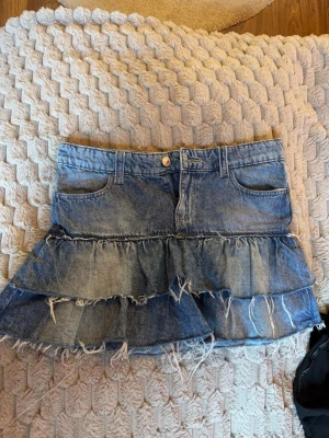 Blå jeanskjol med volanger - Säljer på grund av att jag inte använder den. Det är en blå jeanskjol med två lager volanger och råa, fransiga kanter. Kjolen har klassiska fickor fram och bak samt bälteshällor. Materialet är denim och kjolen har en cool, avslappnad vibe.