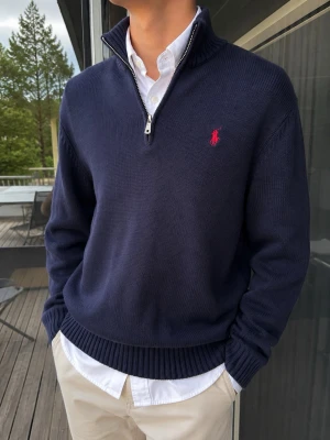 Polo Ralph Lauren Half Zip – Marinblå, Storlek M - Klassisk Polo Ralph Lauren half zip i stilren marinblå färg med den ikoniska röda hästloggan broderad på bröstet. Stickad modell med ribbad krage, muddar och nederkant samt silverfärgad dragkedja. En tidlös och preppy tröja som passar perfekt över en skjorta eller T-shirt. Storlek M. Mycket fint skick 