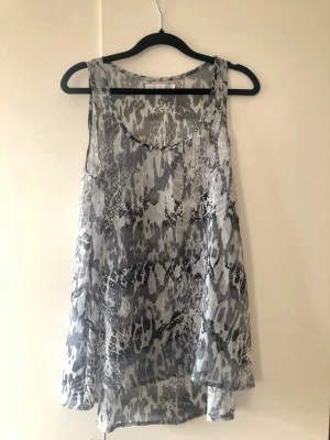 Mönstrat Skirt Linne från ONLY – Stl 34 - Luftigt och skirt mönstrat linne från ONLY i nyanser av grått och svart. Snygg, lite längre modell med fint fall som är något längre baktill. Storlek: 34 (XS). Material: Material-/tvättlapp är avklippt, men tyget känns som tunn polyester/chiffong. Obs! Det finns en horisontell tråddragning/repa i tyget (se bild), men tack vare det levande mönstret syns den inte tydligt vid användning. I övrigt i fint skick!