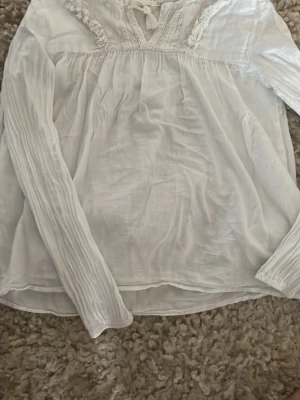 Vit boho-blus med spetsdetaljer - Säljer en luftig vit blus i boho-stil med långa ärmar. Blusen har fina spets- och broderidetaljer runt ringningen samt en liten knytning med tofs framtill. Materialet är tunt och lätt, perfekt för en avslappnad look.