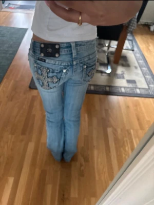 Ljusblå jeans med korsdetaljer - Säljer ett par ljusblå miss me jeans bootcut och snygga korsdetaljer i silver och nitar på bakfickorna. Jeansen har låg midja och dekorativa sömmar, perfekta för dig som vill sticka ut lite extra. Sytt bootcut själv! 