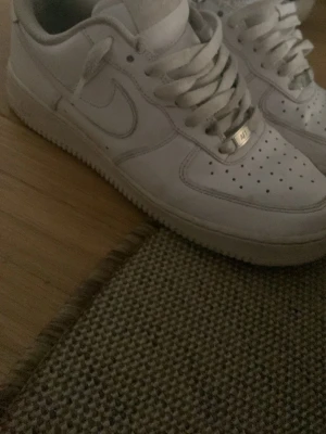 Vita Nike Air Force 1 sneakers - Klassiska vita Nike Air Force 1 sneakers med lågt skaft. Skorna har perforerad tå, vita skosnören och en robust sula. Tillverkade i läder med diskret Nike-logga på sidan och metallbricka på snörningen. Perfekta för en clean och stilren look.
