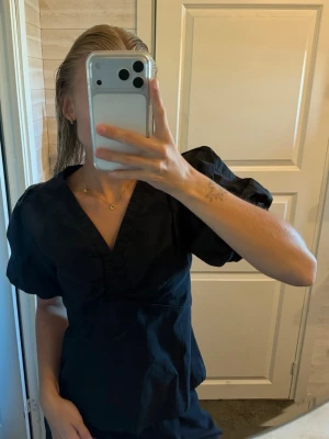 Svart blus med puffärm och v-ringning - Snygg svart blus med puffiga kortärmar och djup v-ringning. Blusen har dekorativa sömmar och lätt glansigt tyg som ger en elegant känsla. Perfekt att styla med jeans eller kjol för en trendig look.