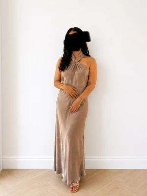 Ny oanvänd satin långklänning med halterneck - Säljer en elegant beige långklänning i ett glansigt material med halterneck och omlottdetalj vid halsen. Klänningen har en mjuk, följsam passform och är ärmlös, perfekt för dig som gillar stilrena och tidlösa plagg.