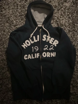 Mörkblå Hollister hoodie med dragkedja - Mörkblå hoodie från Hollister med vit text 'Hollister 1922 California' på bröstet. Tröjan har dragkedja framtill, två fickor och en justerbar huva med vita snören. Insidan av huvan är ljusgrå. Perfekt för en avslappnad streetstyle. Jag hittade ingen storlek på den här har förmodligen åkt bort men 61 cm lång är den om man bortser från luvan och 49 cm bred. Den passar bra till mig som är 172 cm lång