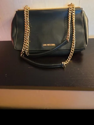 Svart axelväska från Love Moschino - Snygg svart axelväska från Love Moschino med guldfärgade kedjehandtag och detaljer. Väskan har en rektangulär form och ett lock med präglad text och mönster i guld. Tillverkad i slätt konstläder som ger en lyxig känsla.