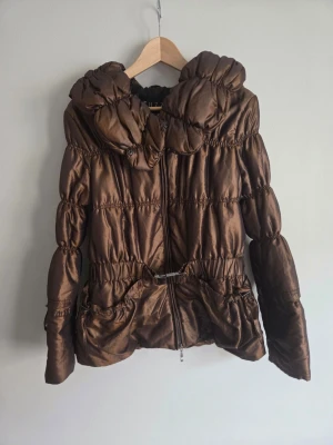 🤎 Lyxig kopparbrun pufferjacka från Tuzzi 🤎 - En otroligt fin och feminin jacka i en vacker mörkbrun/kopparbrun nyans med glansig finish. Jackan har en mysig stor puffkrage, dragkedja framtill och ett snyggt spänne i midjan som skapar en fin och feminin silhuett. En perfekt höstjacka för dig som vill ha en varm men samtidigt stilren och trendig look 🍂 Storlek: Märkt storlek 40, men upplevs mindre i storlek och passar enligt mig en som brukar ha S eller S/M bättre beroende på önskad passform. Mått kan fixas om det önskas. Kolla sista bilden 