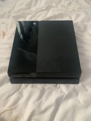 PlayStation 4 spelkonsol med handkontroll - Säljer en svart PlayStation 4 spelkonsol med tillhörande svart DualShock 4 handkontroll. Konsolen har en stilren, rektangulär design med både matt och blank finish. Perfekt för gaming och streaming av film och serier.