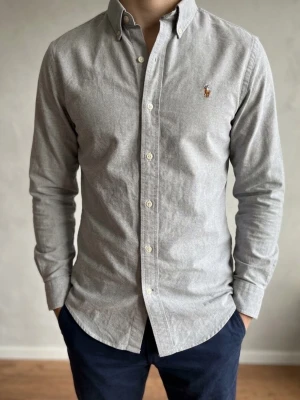 Grå skjorta från Ralph Lauren - Klassisk grå skjorta från Ralph Lauren med button-down krage och broderad logga på bröstet. Skjortan har långa ärmar, vita knappar och är tillverkad i mjuk bomull. Perfekt för en clean och stilren look. Storlek S. Snabb Leverans!