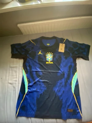 Brasilien borta tröja VM 2026 - Snygg blå Brasilien matchtröja från Nike med gula detaljer och klubbmärke på bröstet. Tröjan har korta ärmar, rund hals och är tillverkad i ett lätt, ventilerande material. Perfekt för fotbollsträning eller att bära till vardags.
