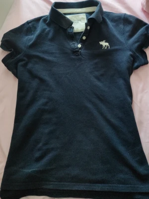 Mörkblå pikétröja med vit logga - Klassisk mörkblå pikétröja från Abercrombie & Fitch med vit älglogga broderad på bröstet. Tröjan har korta ärmar, krage och knappar framtill. Tillverkad i mjukt bomullsmaterial för en bekväm känsla.