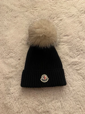 ÄKTA Moncler mössa - Snygg svart ribbstickad mössa från Moncler med stor beige pälsboll på toppen. Mössan har uppvikt kant och Moncler-logga framtill. (Storlek M men är väldigt liten i storlek!!)💗💗💗 finns en likadan mössa fast med svart pälsboll - sista bilden!! (Går även att köpa den) 