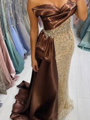 Brun och champagnefärgad långklänning - Elegant långklänning med axelbandslös design i glansig brun satin och champagnefärgad nederdel täckt av paljetter. Klänningen har en dramatisk volangdetalj och en lång släp, perfekt för speciella tillfällen.