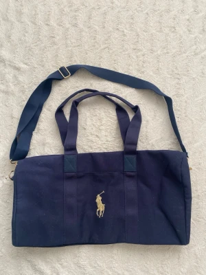 Ralph Lauren duffelbag aldrig använd - Snygg mörkblå axelväska i bomullscanvas med justerbar axelrem som går att ta av och två handtag. Väskan har en guldfärgad dragkedja och en broderad Polo Ralph Lauren-logga i guld på framsidan. 