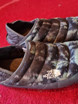 The North Face vadderade tofflor - Säljer ett par vadderade tofflor från The North Face i en cool tie-dye färgskala med blå, svart och grå toner. Tofflorna har en quiltad design och är perfekta för att hålla fötterna varma inomhus. Mjukt innerfoder och slitstark sula.