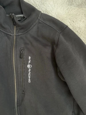 Svart kofta från Sail Racing - Svart kofta från Sail Racing med hög krage och hel dragkedja framtill för män. Koftan har en bröstficka med dragkedja och logga på vänster sida. Tillverkad i ett mjukt material som passar perfekt till kyligare dagar.