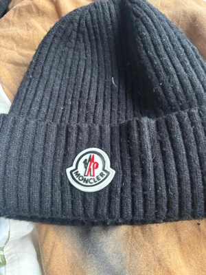 Svart ribbstickad mössa från Moncler - Snygg svart ribbstickad mössa från Moncler med klassisk Moncler-logga framtill. Mössan har uppvikt kant och är tillverkad i ett mjukt, värmande material som passar perfekt för kyliga dagar. Enkel men stilren design som funkar till det mesta.
