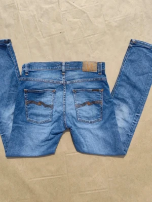 Nudie jeans grim tim - Snygga blå slim fit jeans med slitningar och tvättade detaljer. Klassisk femficksmodell med knappgylf och dekorativa sömmar på bakfickorna. Jeansen har en modern passform och är perfekta för en avslappnad stil. 32x32